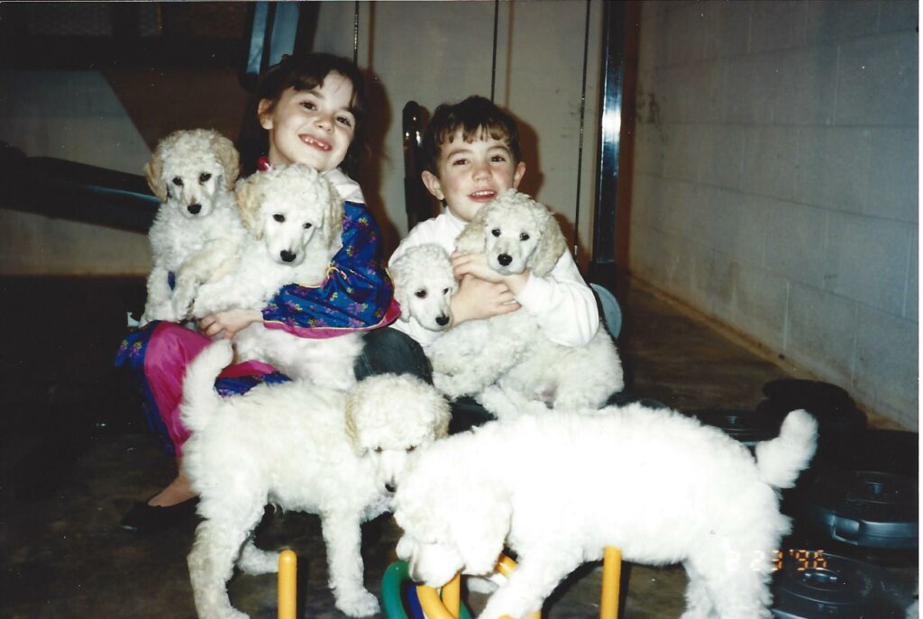 Two smiling children sit on a concrete floor indoors, each holding fluffy white puppies. Three more puppies stand around them, playing. The background includes a treadmill and a block wall.