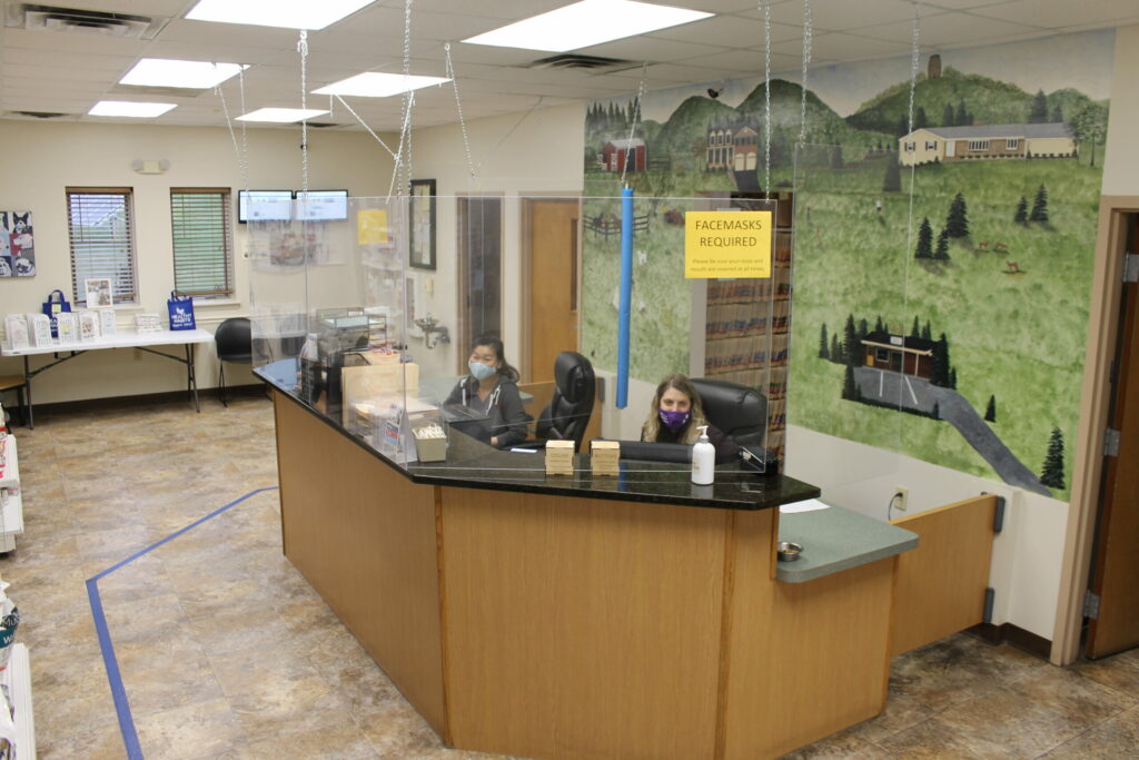 A reception area with two masked staff behind a glass partition, a "Facemasks Required" sign, and a mural of buildings and trees on the wall in the background. Hand sanitizer sits on the counter.