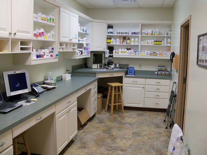 A pharmacy workspace with white cabinets, shelves filled with medicine bottles, a computer on the counter, a microscope, a wooden stool, and various pharmacy supplies neatly organized throughout the room.