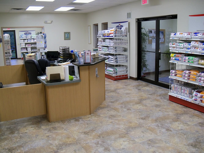 A clean, modern veterinary clinic reception area with a front desk, office chairs, shelves stocked with pet food, and a glass entrance door. The space has tiled flooring and bright fluorescent lighting.