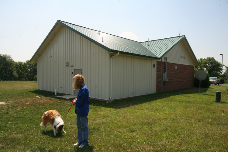 A person in a blue jacket holds a dog on a leash outside a beige building with a green metal roof, standing on grassy ground under a clear sky.